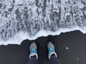 Legs of a person wearing blue and grey hiking boots, one of the coolest outdoor gifts for her. The image is taken on a black sand beach, and the water is coming very close to the hiking boots.