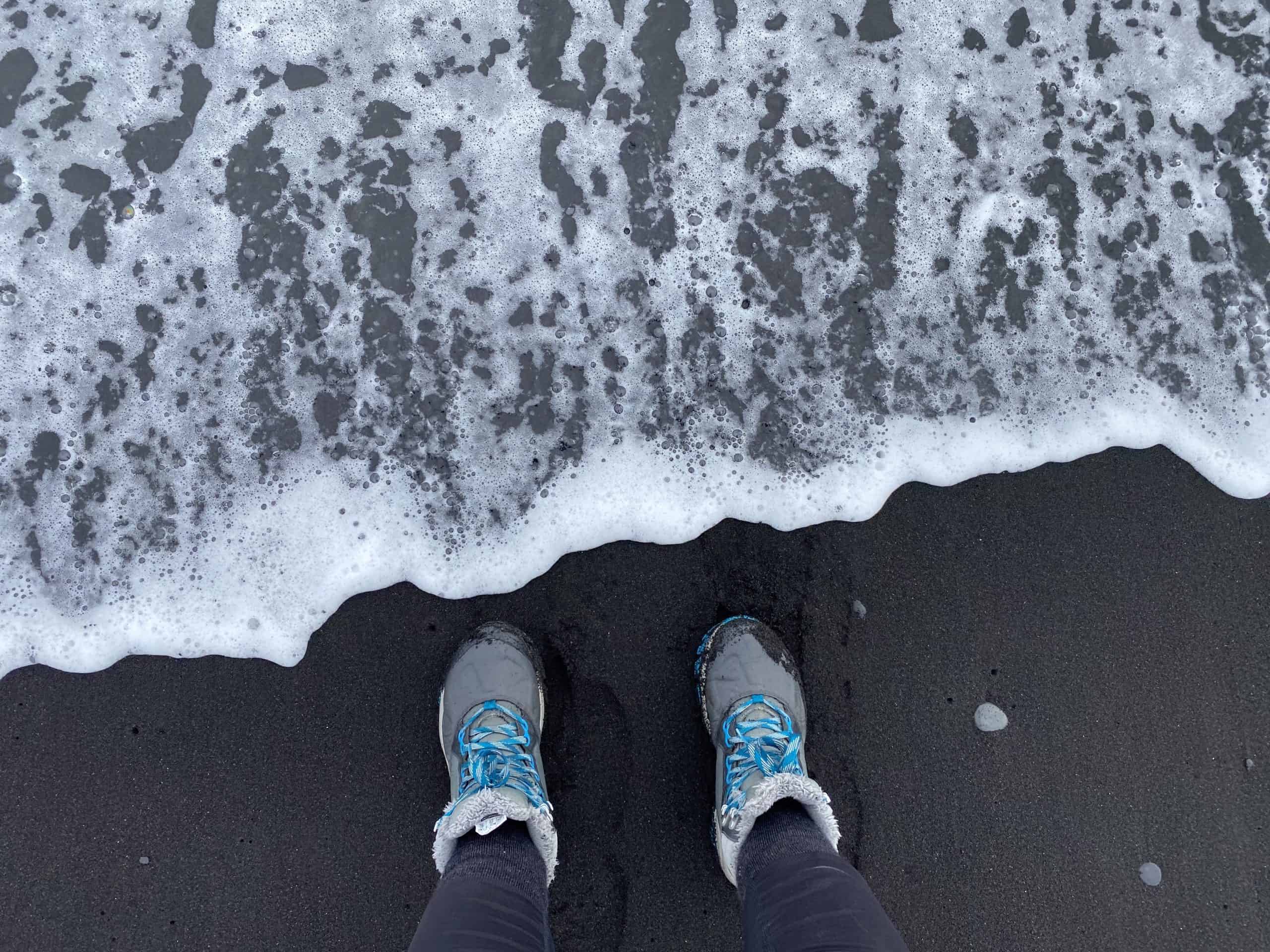 Legs of a person wearing blue and grey hiking boots, one of the coolest outdoor gifts for her. The image is taken on a black sand beach, and the water is coming very close to the hiking boots.
